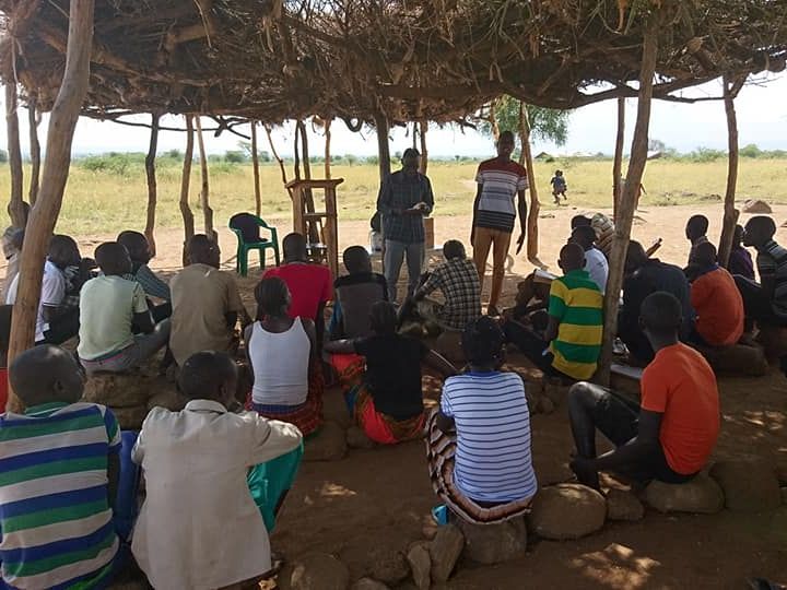 A class meeting outdoors under a thatched shelter